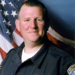 Image depicts an official photo of Officer Andrew Stevens with a flag in the background.